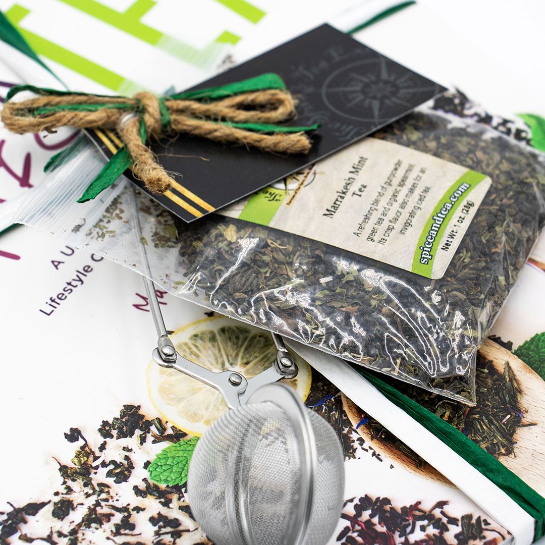 The Cancer Hates Tea Book & Bundle - Marrakesh Mint sits beside a mesh infuser on a magazine, with loose leaves and a lemon slice in the background, emphasizing the link between tea and wellness.