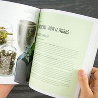A person holds open the Cancer Hates Tea Book & Bundle - Ginger Turmeric, displaying a chapter titled Cancer 101—How It Works and glass jars of dried herbs, connecting tea and wellness against a dark, textured background.