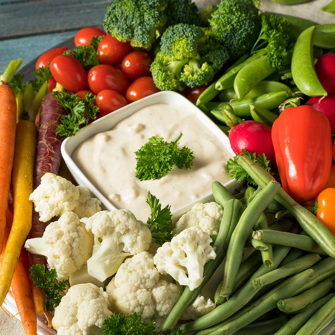Assorted vegetables including carrots, broccoli, tomatoes, and cauliflower with a dip in the center on a wooden surface.