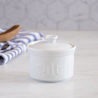 The White Ceramic Salt Cellar, labeled "salt," rests on a marble countertop with a striped towel and wooden spoon nearby, all set against a hexagonal tile backsplash—an ideal, stylish solution for artisan salt storage in your kitchen.
