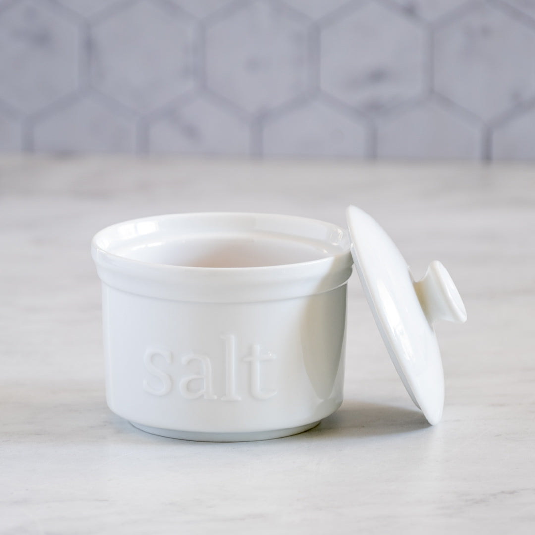 The White Ceramic Salt Cellar, with "salt" embossed on its front, sits on a marble countertop with its lid set aside. Ideal for storing artisan salts, it contrasts beautifully against a light gray hexagon tile background.