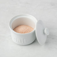 The White Ceramic Salt Cellar features a matching lid and holds pink artisan salts, making it a stylish and practical storage solution for your kitchen.