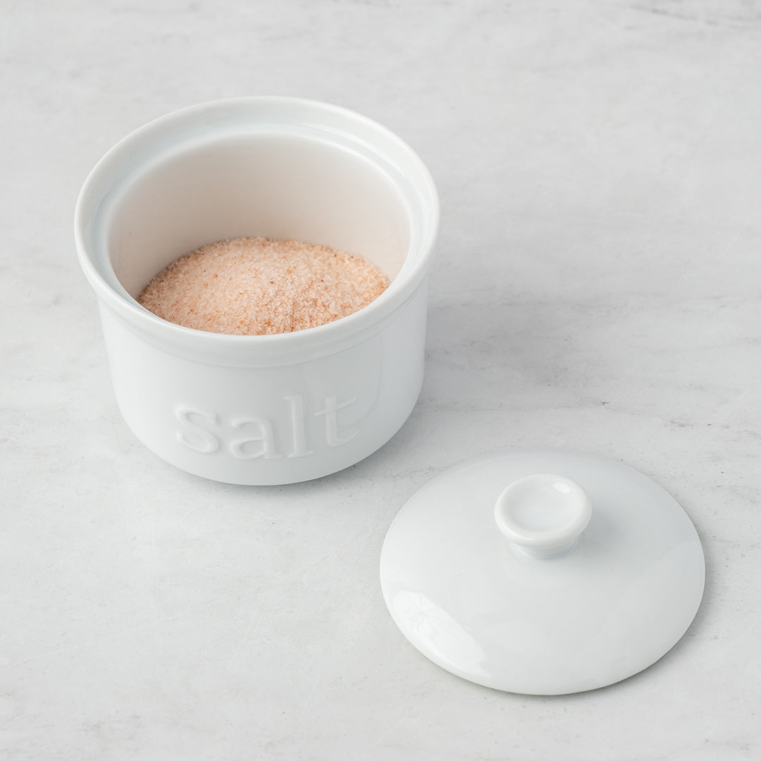 The White Ceramic Salt Cellar holds pink Himalayan salt, with its matching lid set beside it on a light gray marble surface—an elegant option for stylish kitchen storage.