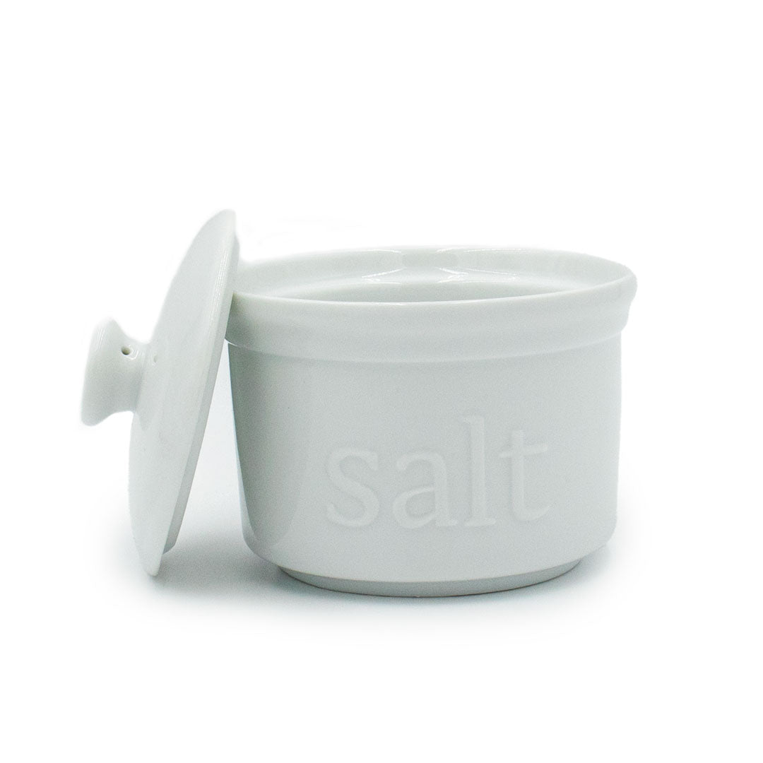 The White Ceramic Salt Cellar is ideal for artisan salts, featuring "salt" embossed on the front and a matching lid resting beside it. This stylish white ceramic container is perfect for kitchen storage and display.