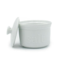 The White Ceramic Salt Cellar is ideal for artisan salts, featuring "salt" embossed on the front and a matching lid resting beside it. This stylish white ceramic container is perfect for kitchen storage and display.