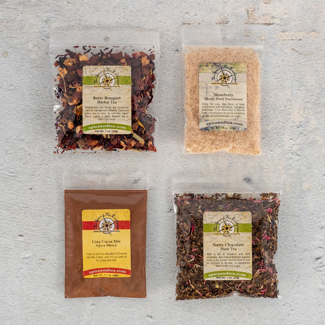 The Chocolate-Dipped Strawberry Tea Sampler features four clear bags on a light gray surface, each with berry bouquet herbal tea, strawberry monk fruit sweetener, cozy cocoa mix, and nutty chocolate mate for unique drink blends.