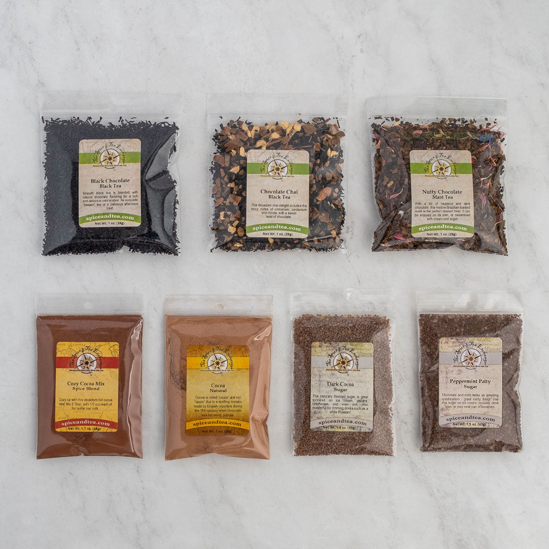 Seven clear plastic bags from the Chocolate Lover's Gift Box display assorted loose leaf teas and powders on a light surface, with labels for flavors like Black Chocolate, chocolate chai, Nutty Chocolate, and rich cocoa blends.