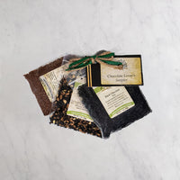 A packaged chocolate sampler gift set tied with a green ribbon, placed on a light marble surface. The set includes three small bags of tea, with a card reading 'Chocolate Lover's Sampler'.