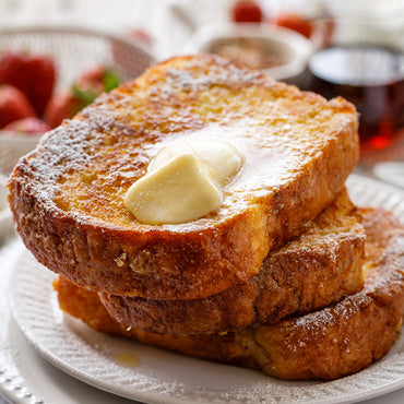 Stack of French toast with butter on a white plate, surrounded by fruits and syrup.