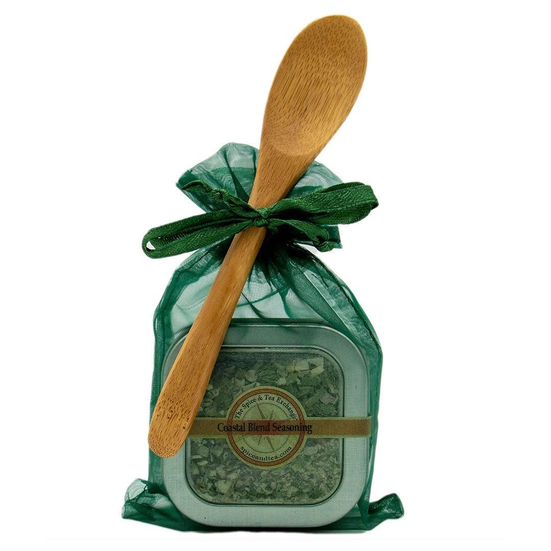 The Coastal Blend Seasoning - Spice Tin with Spoon comes in a green mesh gift bag, tied with a green ribbon, and includes a wooden spoon attached diagonally across the front—perfect for seasoning seafood.
