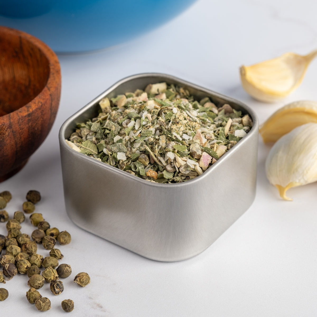 A square metal tin of Coastal Blend Seasoning rests on a white surface, surrounded by garlic cloves, black peppercorns, and a wooden bowl—an all-purpose mix ideal for adding a cool, coastal flavor to any dish.