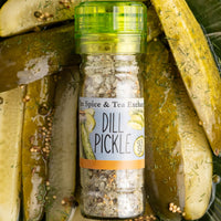 A green-capped bottle of Dill Pickle Sea Salt Seasoning from The Spice & Tea Exchange, perfect as a pickle grinder, sits among sliced dill pickles, showing off its tangy sea salt and visible spices inside.