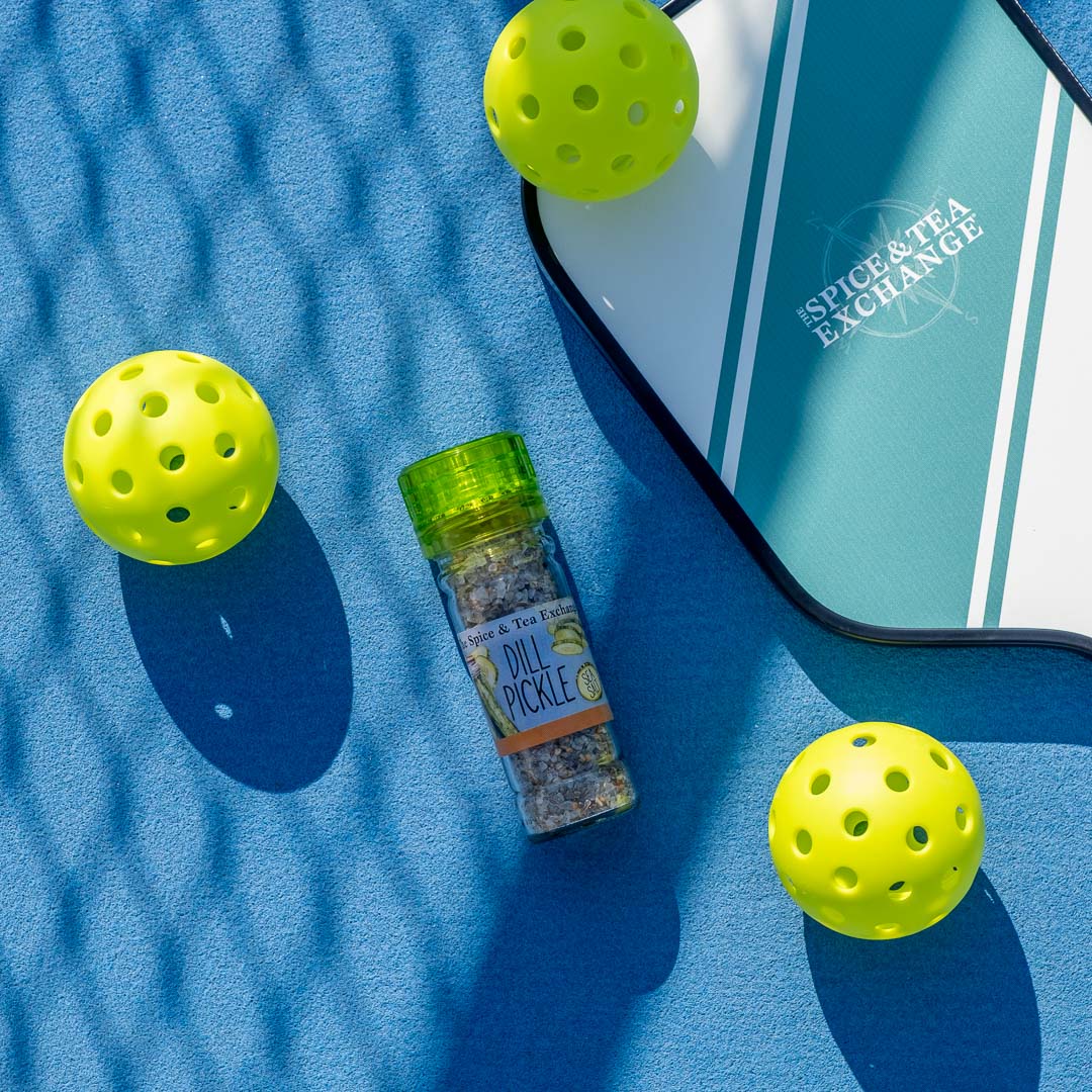 A container of Dill Pickle Sea Salt Seasoning rests on a blue surface beside a pickleball paddle and three yellow pickleballs beneath a mesh shadow.