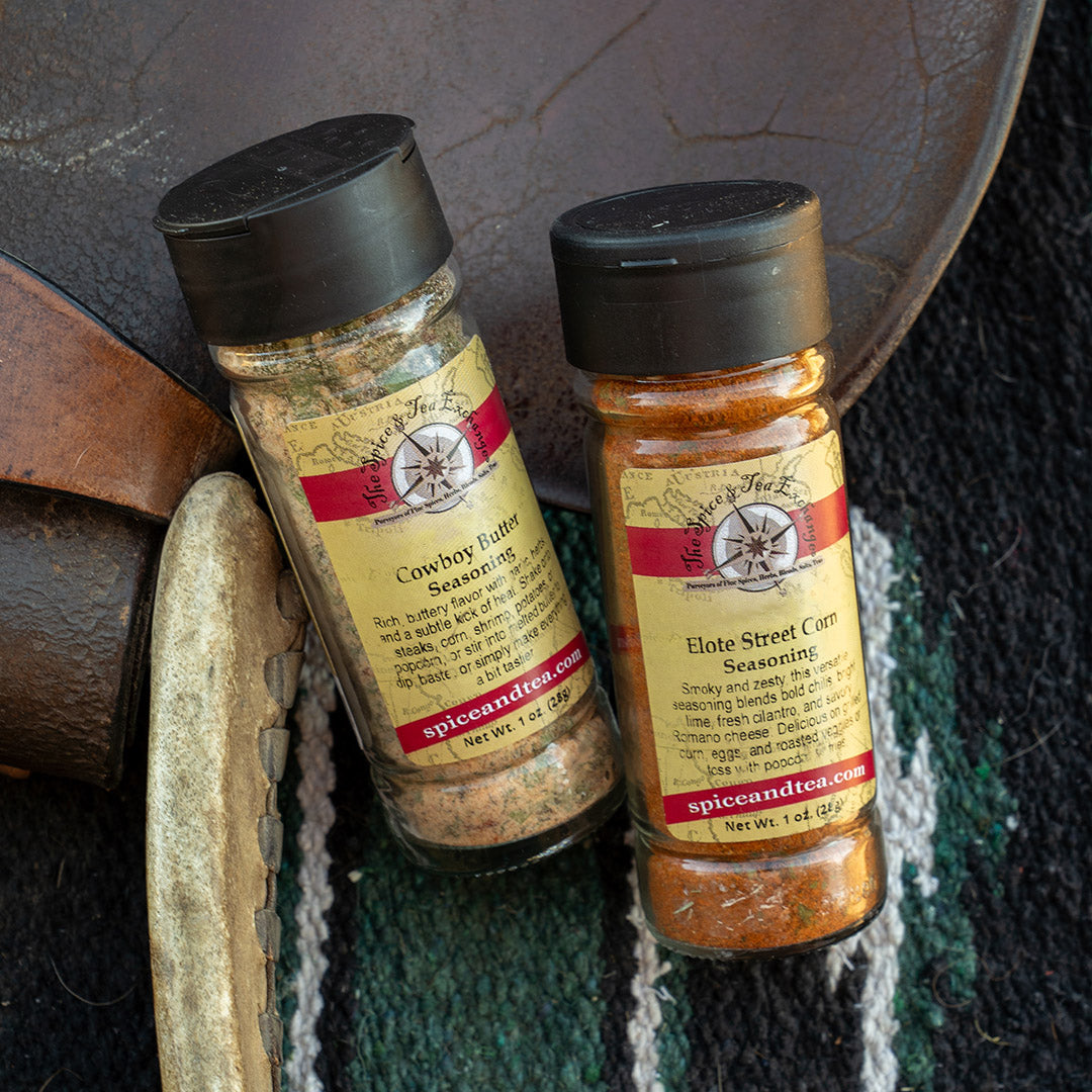 Two spice seasoning bottles labeled Spiceland's 'Cowboy Rubbed Seasoning' and 'Elote Street Corn Seasoning' resting on a rustic leather and woven textile background.