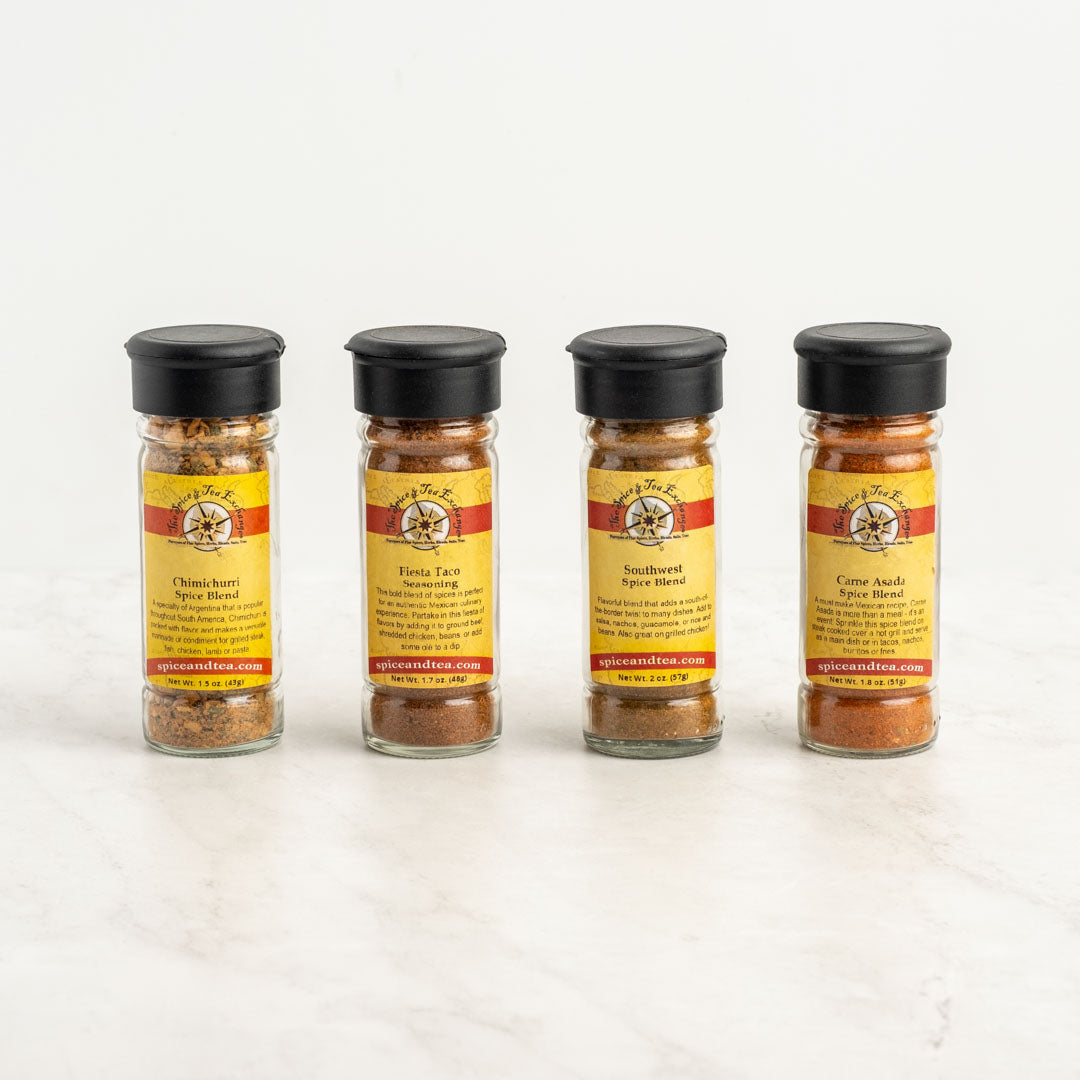 The Fiesta Flavors 4-Pack Gift Box includes four spice jars with black lids and yellow-red striped labels, each containing a different Latin American seasoning from Spice and Tea Co., arranged in a row on a white background.