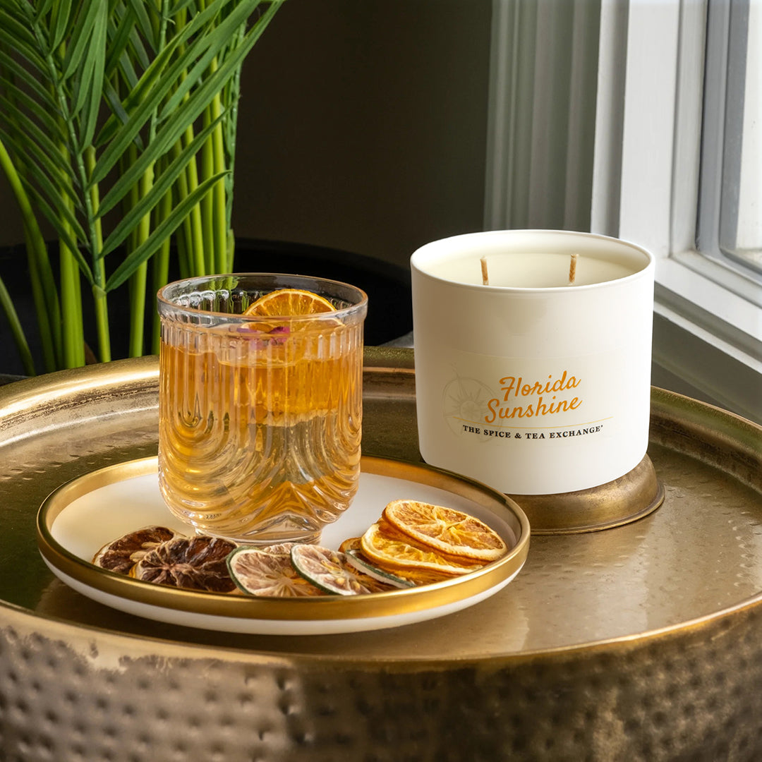 A glass of amber-hued Florida Sunshine White Tea with a dried orange slice, a matching white candle, and assorted dried citrus slices on a gold tray by the window beside a green plant.