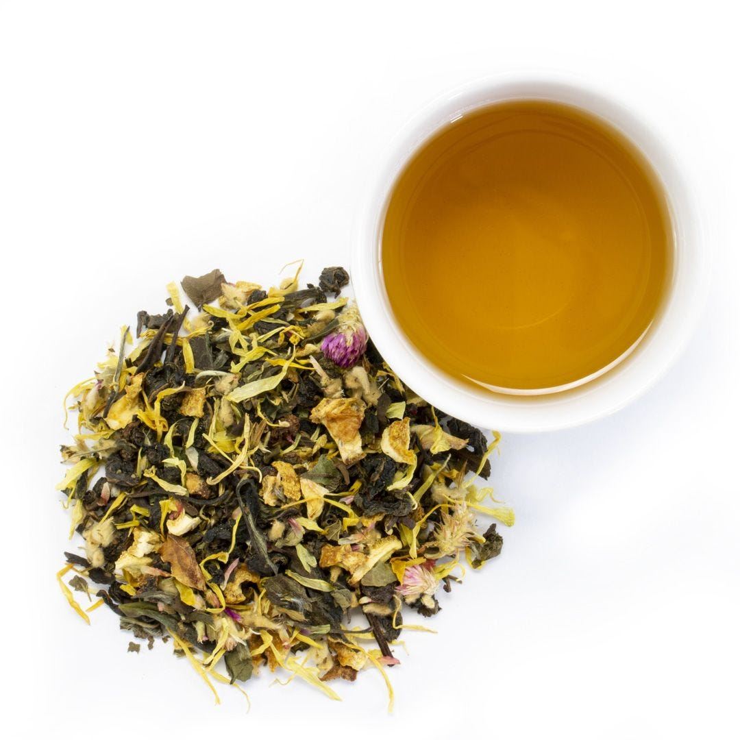 A white cup of Florida Sunshine White Tea sits beside a pile of loose leaf tea with green leaves, yellow petals, and dried flower buds on a white background.