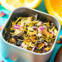 A close-up of a metal container filled with Florida Sunshine White Tea, a loose leaf blend with dried flowers, fruit peels, and leaves. Fresh orange slices highlight this floral and citrus tea on a bright blue surface.
