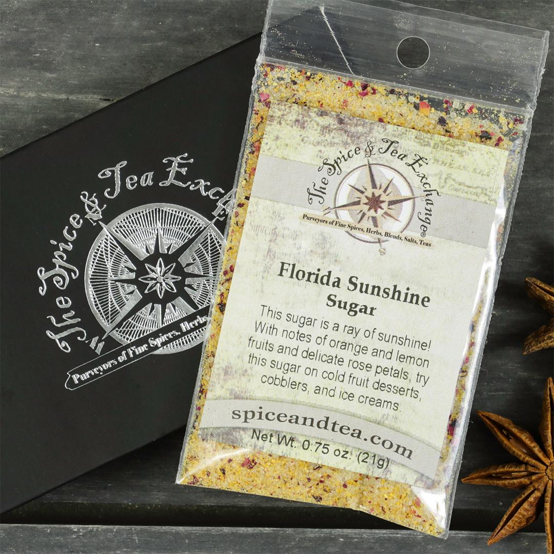 A packet labeled "Florida Sunshine Sugar - Barter Box," a citrus sugar and flower petal mix, rests on a gray tray next to a black Spice & Tea Exchange box with star anise accents, offering a unique gift choice.