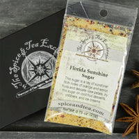 A packet labeled "Florida Sunshine Sugar - Barter Box," a citrus sugar and flower petal mix, rests on a gray tray next to a black Spice & Tea Exchange box with star anise accents, offering a unique gift choice.