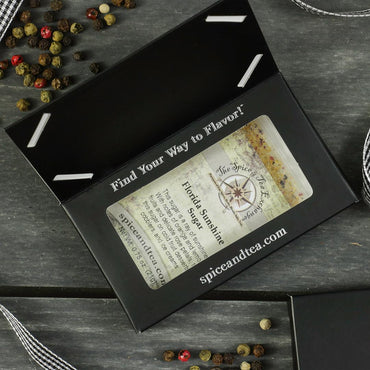 A black box with a clear window showcases the Florida Sunshine Sugar - Barter Box from The Spice & Tea Exchange, accompanied by colorful peppercorns and a striped ribbon on a gray wooden surface—a unique citrus sugar gift.