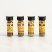 Four spice jars with black lids are lined up on a white surface. The Garlic Lover's 4-Pack Gift Box includes: Smoky Black Garlic, Black Truffle Garlic, Viki’s Garlic Fix, and Garlic n Herb.