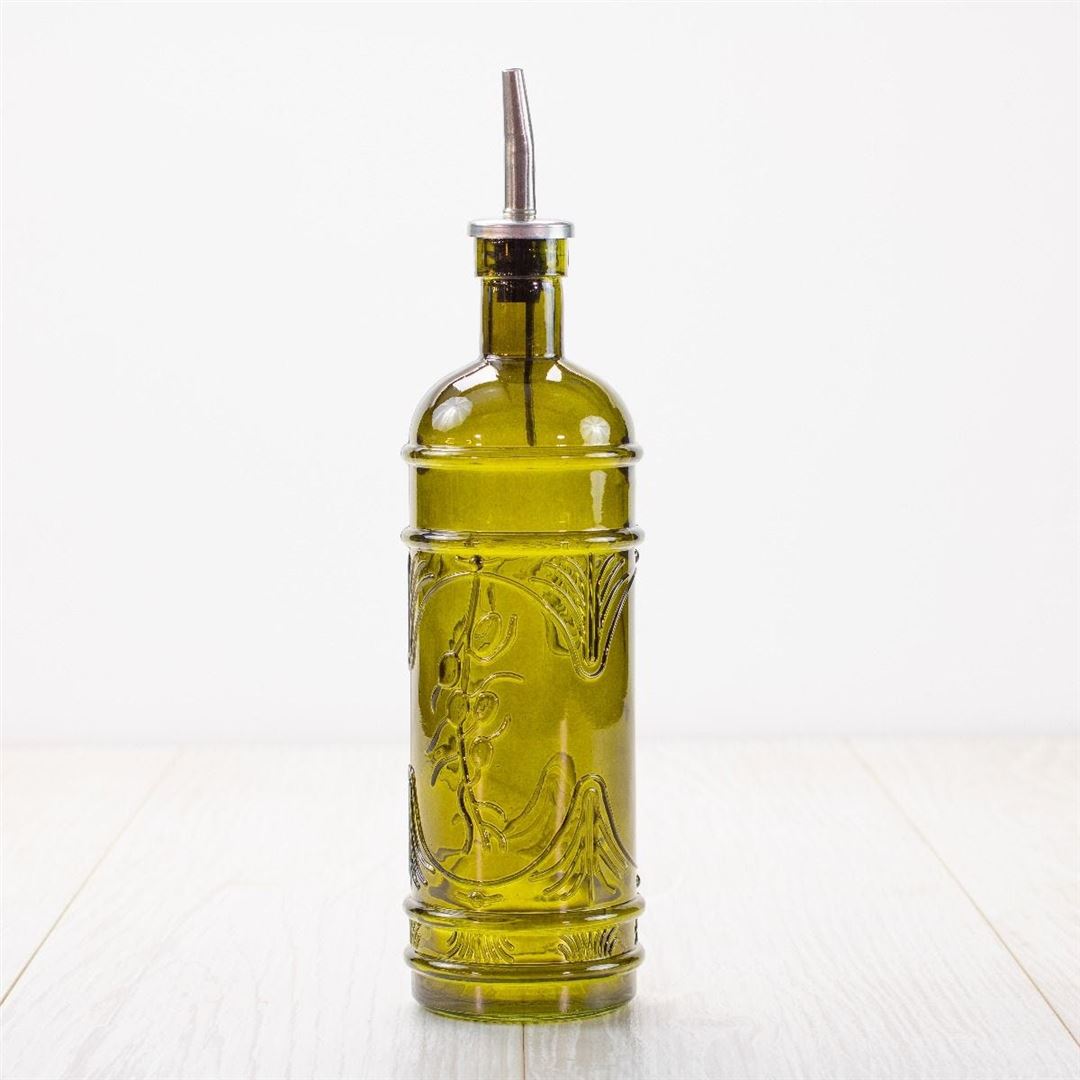 The Olive Glass Oil Jar, made of green glass with an embossed design and a metal pour spout, sits on a light wooden surface against a plain white background.