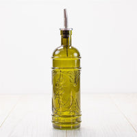 The Olive Glass Oil Jar, made of green glass with an embossed design and a metal pour spout, sits on a light wooden surface against a plain white background.