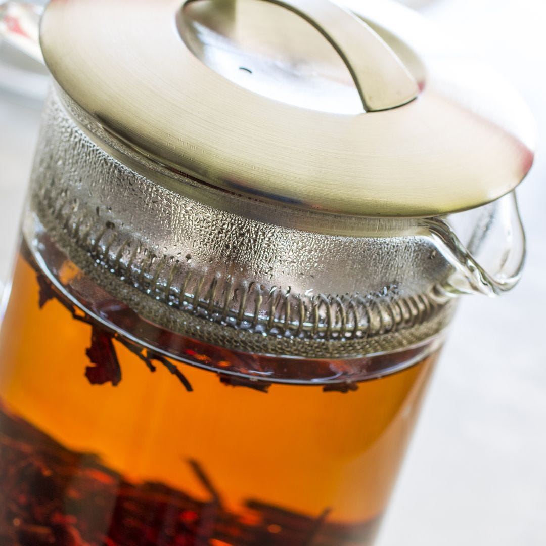 A close-up of the Glass Pitcher with Filter, featuring a metal lid and stainless steel strainer, filled with amber loose-leaf tea and visible tea leaves inside, with steam condensing on the lid.