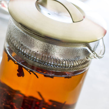 A close-up of the Glass Pitcher with Filter, featuring a metal lid and stainless steel strainer, filled with amber loose-leaf tea and visible tea leaves inside, with steam condensing on the lid.