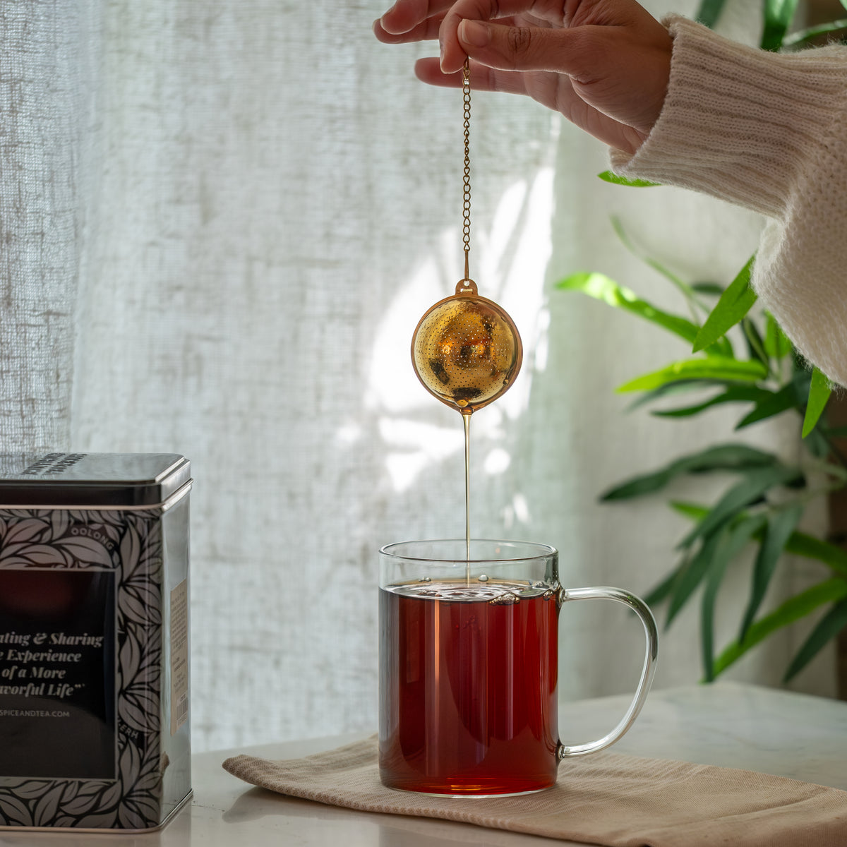 A hand holds the Gold Ball Infuser - Small above a glass mug of tea; sunlight streams through a window onto a green plant, with a decorative tea tin nearby on a beige cloth.