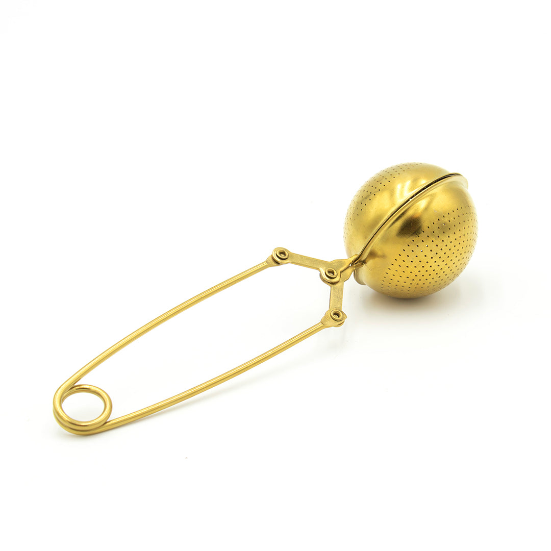 The Gold Scissor Infuser is a metal, gold tea infuser with a round mesh ball and long handle, featuring a scissor design ideal for steeping loose tea leaves.