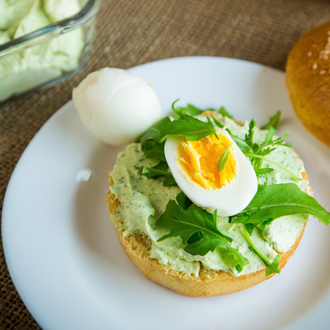 Bagel with avocado spread, boiled egg, and arugula on a white plate.