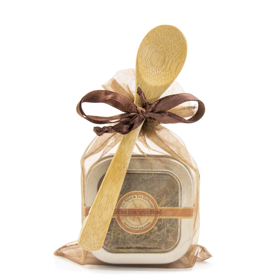 The Grilled Fish Spice Blend - Spice Tin with Spoon comes in a small metal tin with a clear lid, wrapped in a sheer gold organza bag tied with a brown ribbon, and topped with an attached wooden spoon.