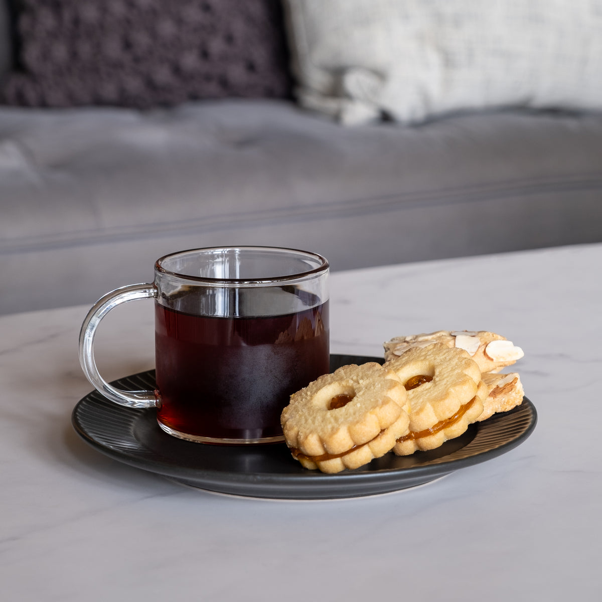 A glass mug of Hazelnut Cookie Tea and three assorted cookies sit on a black plate atop a white marble table, with a grey sofa and cushions in the background.