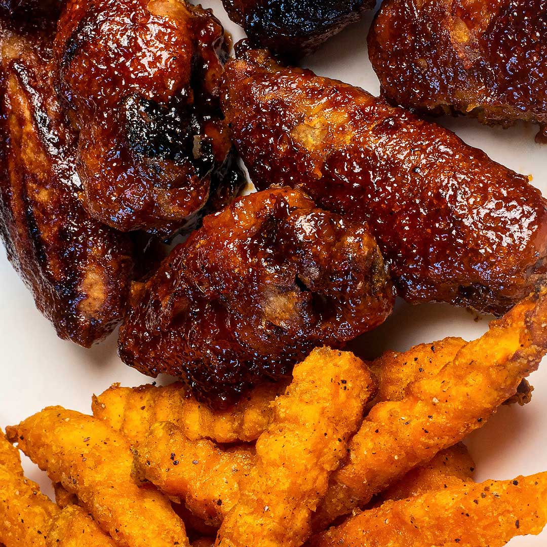 Close-up of glazed barbecue-style chicken wings piled on a white plate, with golden-orange fried tenders or strips in the foreground.