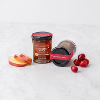 Two Cranberry Apple Jam Honey Jars, one upright and one lying down, are displayed on a white surface with apple slices and whole cranberries.