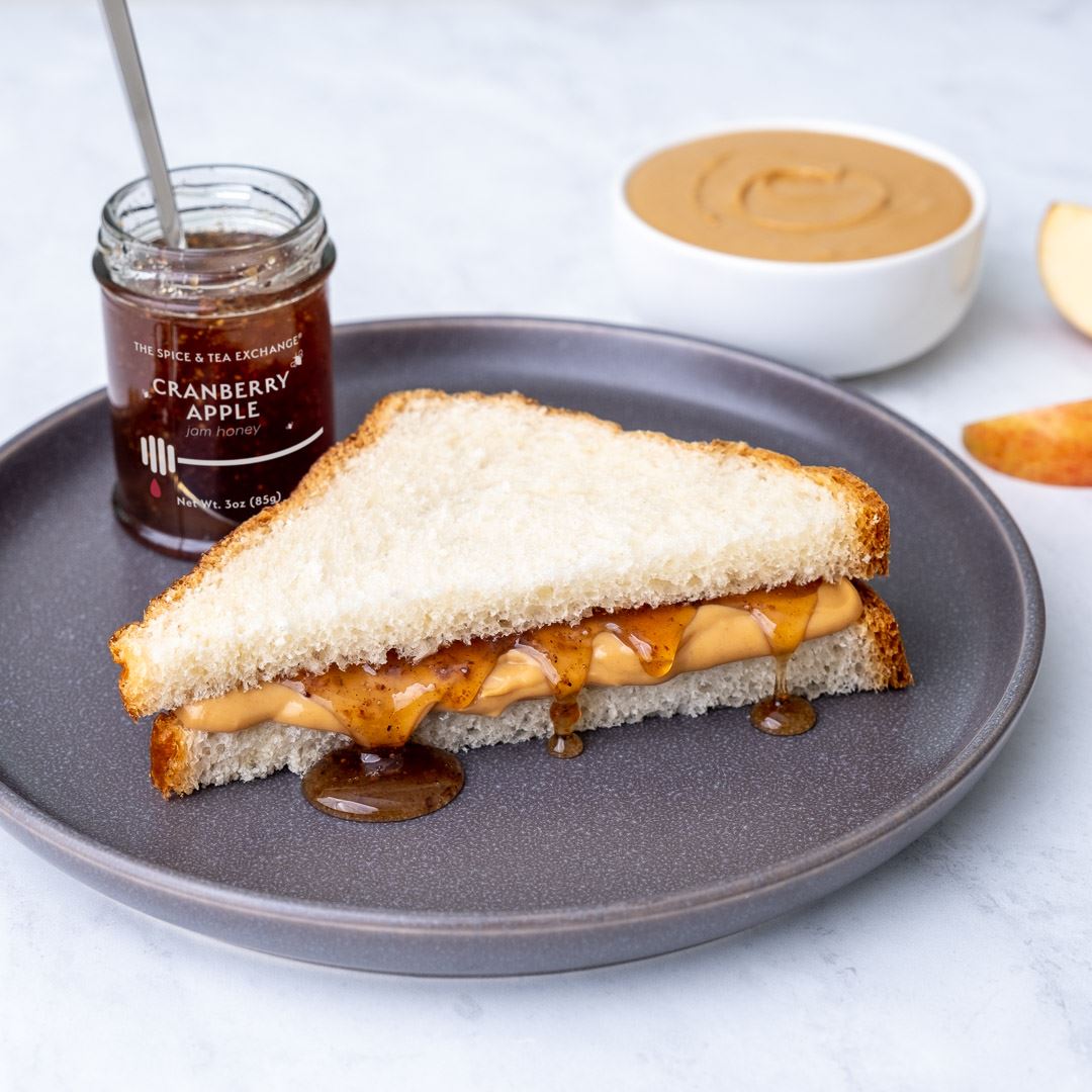 A peanut butter and jelly sandwich on white bread sits on a gray plate. Nearby, a Cranberry Apple Jam Honey Jar, a bowl of peanut butter, and an apple slice complete the tempting scene.