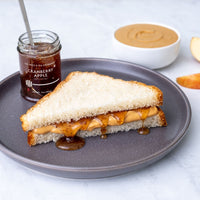 A peanut butter and jelly sandwich on white bread sits on a gray plate. Nearby, a Cranberry Apple Jam Honey Jar, a bowl of peanut butter, and an apple slice complete the tempting scene.