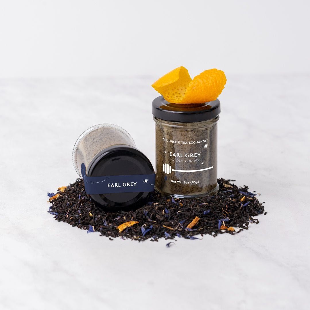 Two Earl Grey Whipped Honey Jars—one upright with orange peel and one lying down—are displayed on a white marble surface, surrounded by tea leaves and blue flower petals.