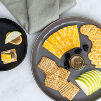 A round tray holds sliced marbled cheese, crackers, green apple slices, blackberries, and an Earl Grey Whipped Honey Jar. On a small plate: a cracker with cheese, honey, and apple. A gray napkin is set nearby.