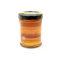 The Himalayan Salted Honey Jar is a 3 oz (85 g) glass jar with a black lid and yellow label showing a honey dipper. Honey drips down the side. Perfect for a cheese board. Background is white.