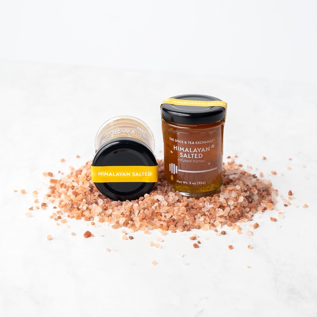 Two Himalayan Salted Honey Jars, one upright and one lying down, are showcased on a bed of pink Himalayan salt crystals—perfect for elevating your next cheese board.