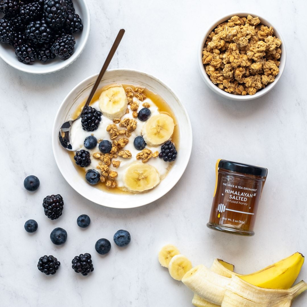 A bowl of yogurt with banana slices, blackberries, blueberries, and granola sits on a white surface. Nearby are a bowl of granola, blackberries, a banana, and a jar of Himalayan Salted Honey.