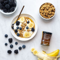 A bowl of yogurt with banana slices, blackberries, blueberries, and granola sits on a white surface. Nearby are a bowl of granola, blackberries, a banana, and a jar of Himalayan Salted Honey.