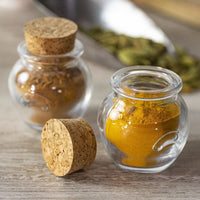 A Glass Jar Honey Pot with Cork sits open on a wooden surface containing ground turmeric, while another holds cinnamon. Cardamom pods are blurred in the background, highlighting the jars’ charm for stylish spice storage.