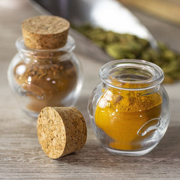 A Glass Jar Honey Pot with Cork sits open on a wooden surface containing ground turmeric, while another holds cinnamon. Cardamom pods are blurred in the background, highlighting the jars’ charm for stylish spice storage.