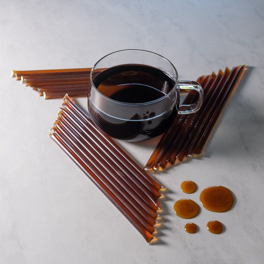 A glass mug of dark coffee sits on a white surface, surrounded by Bourbon Honey Straws and several brown coffee concentrate sticks arranged in a geometric pattern, with drops of coffee nearby.