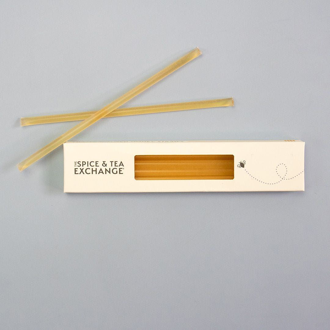 A white box labeled The Spice & Tea Exchange with a clear window displays several Lemon Honey Straws filled with raw natural honey. Two straws are placed diagonally above the box on a plain light gray surface, perfect as a tasty, healthy snack.