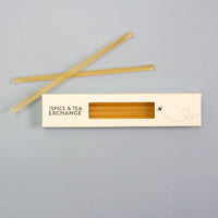 A white box labeled The Spice & Tea Exchange with a clear window displays several Lemon Honey Straws filled with raw natural honey. Two straws are placed diagonally above the box on a plain light gray surface, perfect as a tasty, healthy snack.